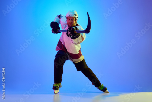 Photography Competitive, motivated man, hockey player posing with stick, in uniform and helmet against gradient blue background in neon light