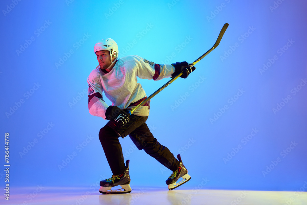 Obraz premium Man, hockey player wearing uniform and helmet, posing with stick against gradient blue background in neon light. Concept of professional sport, competition, game, tournament