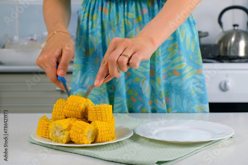 Woman cutting piece of baked corn on plate on kitchen at home to eat, hand close-up. Prepare cook bake dish. Cuisine culinary nutrition domestic food recipe ingredients vegetable. Cooked sweet corn.