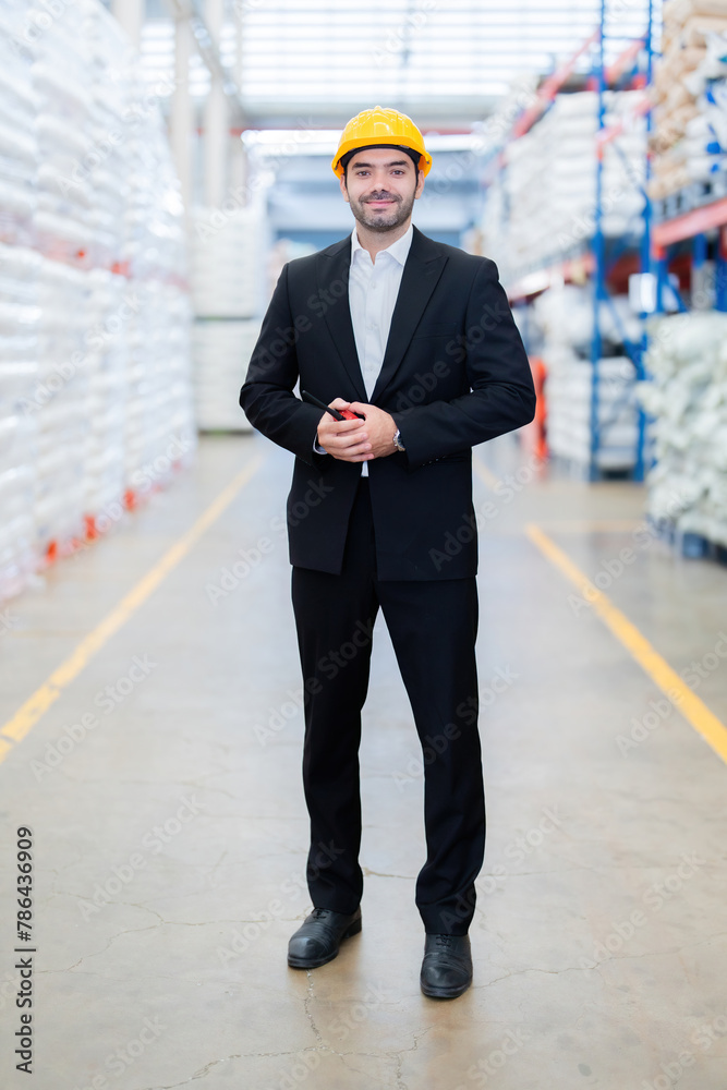 Full body image of a European male engineer, factory owner, holding a walkie-talkie, wearing a helmet and a suit. Look Smart inspects systems in factories that export plastic and steel products. - obrazy, fototapety, plakaty 