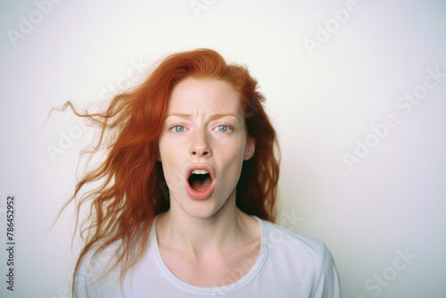 Shocked young woman with ginger hair standing near gray wall