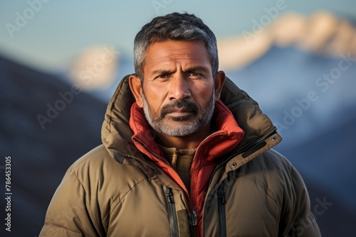 Portrait of a tender indian man in his 50s donning a durable down jacket on backdrop of mountain peaks