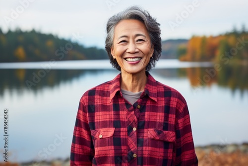 Portrait of a grinning asian woman in her 50s wearing a comfy flannel shirt over serene lakeside view photo