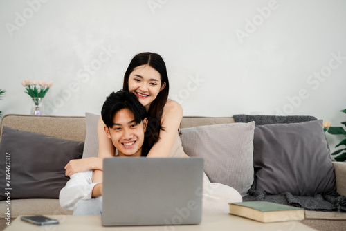 Happy young couple shares a cozy and intimate moment, with the woman embracing the man from behind as they both focus on a laptop screen. © Mongta Studio