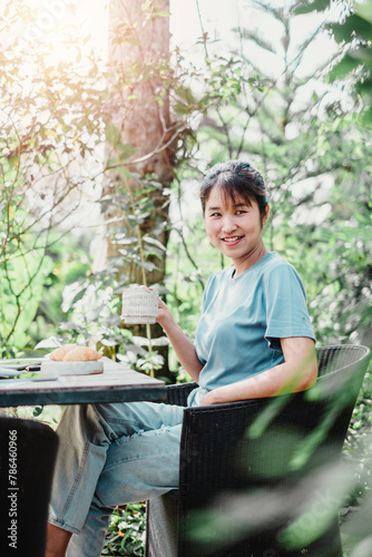 Cheerful woman holds a coffee mug seated in a wicker chair, with green foliage and sunlight filtering through trees behind her.