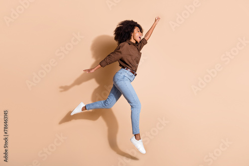 Full size photo of optimistic woman dressed brown shirt jeans jumping look empty space raising fist up isolated on beige color background