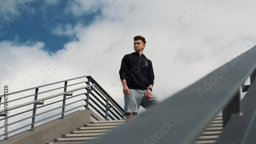 Confident young man in sportswear stands on the stairs and looks around and into the camera. Sport lifestyle