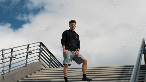 Confident young man in sportswear stands on the stairs and looks around and into the camera. Sport lifestyle