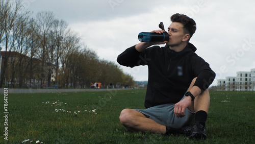 A young man drinks water from a sports bottle. Resting after a run. Sitting in a city park. Healthy Lifestyle