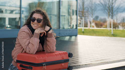 A young woman with a red suitcase is waiting for a transport, a bus outdoors. Smiles dreamily. Urban lifestyle