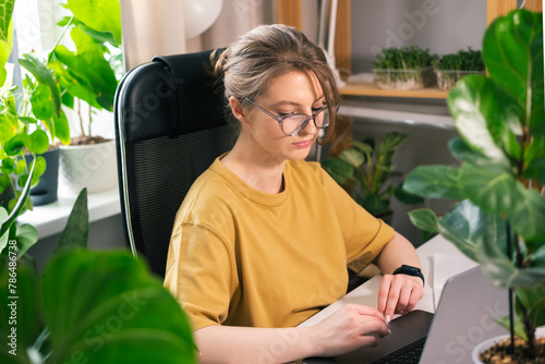 Beautiful young woman working on laptop from home surrounded by indoor plants. Concept of remote work, freelancing, online learning, in the urban jungle.