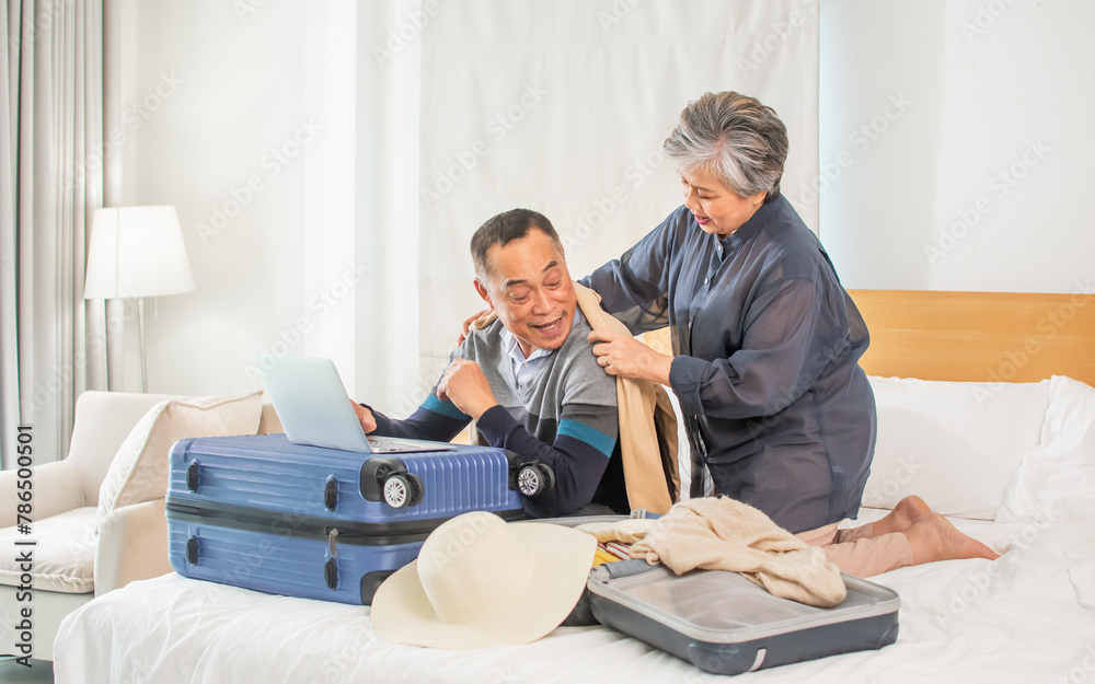 Asian aging sweet couple sitting on bed in bedroom at home, prepare clothes, packing in luggages or baggages for summer travel, smiling with happiness. Tourism, Aging Society, Healthcare concept.