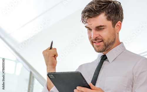 Portrait Caucasian smart handsome businessman wearing formal business clothes, holding, using tablet for working with happy face, standing in indoor modern office or workplace with copy space.
