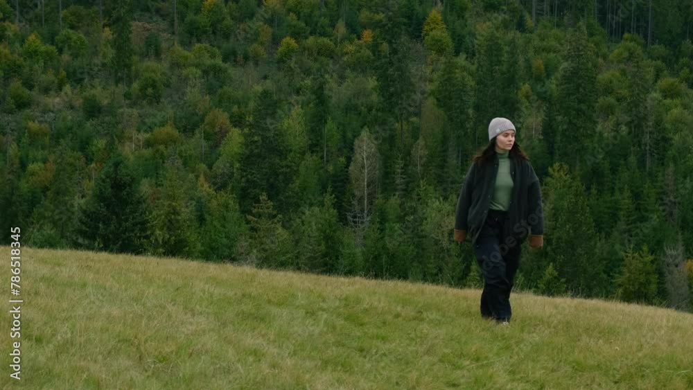 Woman walking on grass observing mountain peaks panorama. Female traveler climbs a hill against the background of a dense mountain forest on cloudy day Woman walking on grass observing mountain peaks panorama. Female traveler climbs a hill against the background of a dense mountain forest on cloudy day