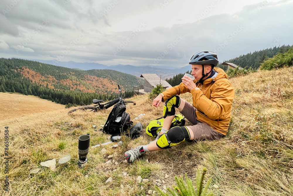 Fototapeta premium Man cyclist drinking a cup with tea, wearing bike helmet, sitting on grass in the mountains, resting after riding electric bike. Mountain e-bike lying on the ground next to him.
