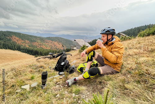 Man cyclist drinking a cup with tea, wearing bike helmet, sitting on grass in the mountains, resting after riding electric bike. Mountain e-bike lying on the ground next to him.