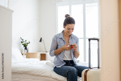 Young woman with a suitcase sitting on bed in hotel room and using phone
