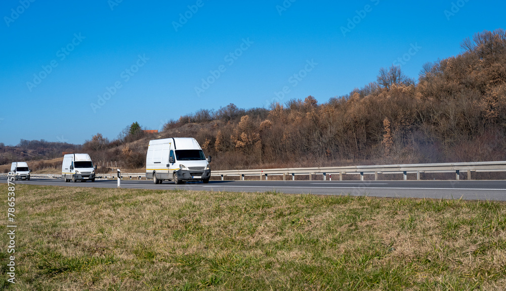 Three white minivans are moving in a convoy on the highway. White delivery van on the highway. White modern delivery small shipment cargo courier van moving fast on motorway road to city urban suburb Three white minivans are moving in a convoy on the highway. White delivery van on the highway. White modern delivery small shipment cargo courier van moving fast on motorway road to city urban suburb