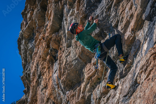 Close-up Caucasian man with black hair. Practicing rock climbing on overhanging wall. Performing athletic movement. With rock wall and blue sky in the background