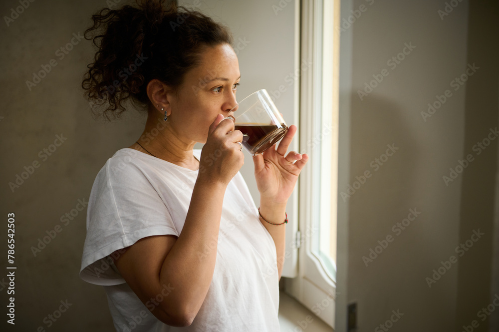 Happy young Latina woman in white pajamas, enjoying her cup of hot drink at home, standing by window overlooking cityscape. Happy young Latina woman in white pajamas, enjoying her cup of hot drink at home, standing by window overlooking cityscape.