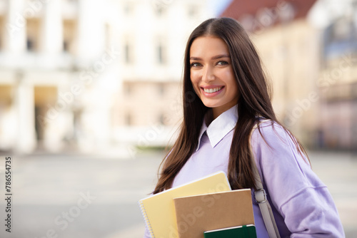 Smiling young woman holding books outdoors at university campus © Prostock-studio