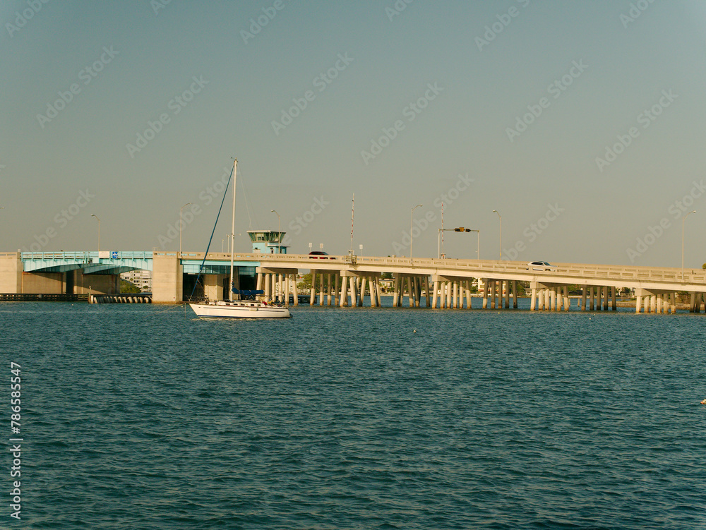 Fototapeta premium Wide View South East from St Pete Beach Community Center towards Corey Causeway drawbridge Boca Ciega Bay with sailboat no mast in blue water near sunset. In Florida. 