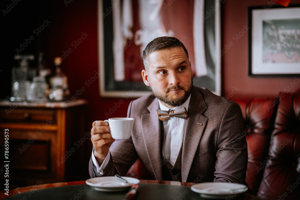 Valmiera, Latvia- July 28, 2024 - A man in a brown suit and patterned bow tie sits at a table, holding a white cup, with a contemplative look, in a vintage style lounge with leather seating. Valmiera, Latvia- July 28, 2024 - A man in a brown suit and patterned bow tie sits at a table, holding a white cup, with a contemplative look, in a vintage style lounge with leather seating.