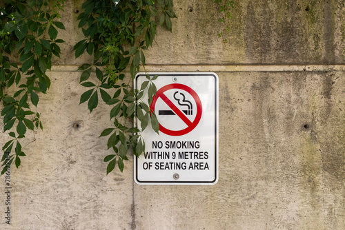 No smoking sign - affixed to a concrete wall - partially obscured by overgrown green leaves. Taken in Toronto, Canada.