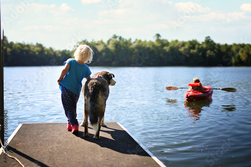 Happy Little Girl And her Big Dog Watching Her Brother Boating in a Kayak on Lake in Woods