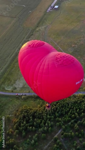 Red aerostat over fields. Romantic hot air balloon in a form of a heart travels in the sky on beautiful nature background at sunset. Top aerial view. Vertical video