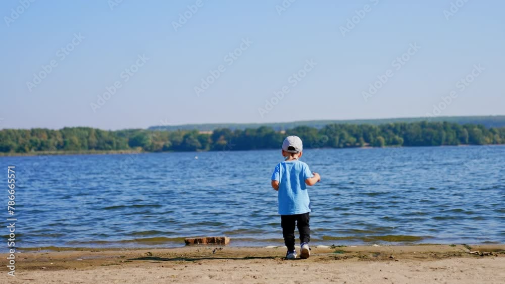 Rear view of a toddler boy heading to the river. Little child walks by the waterfront in summer. Rear view of a toddler boy heading to the river. Little child walks by the waterfront in summer.