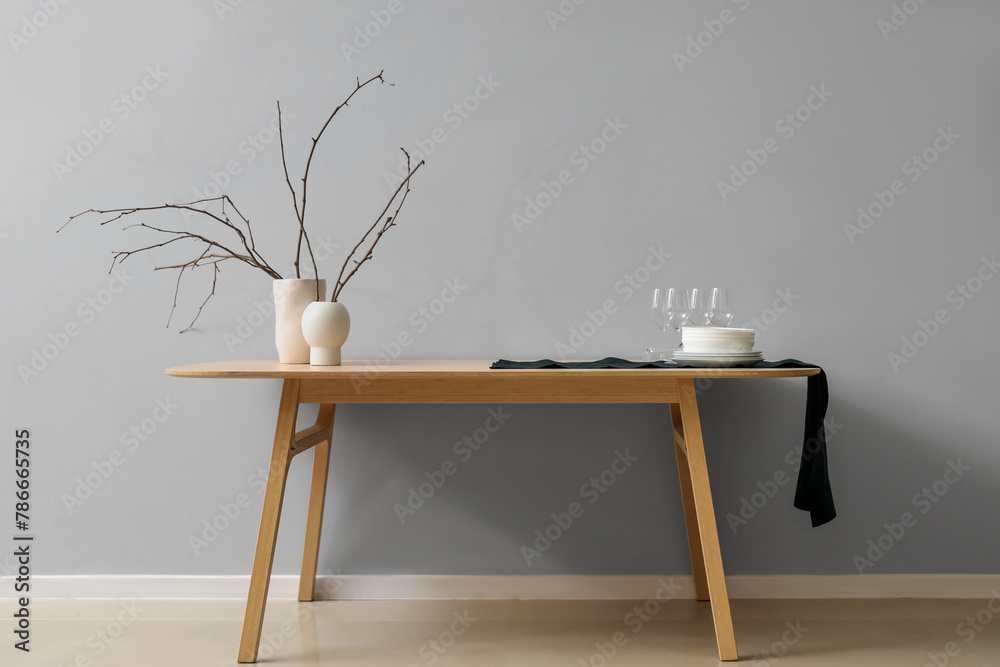 Clean tableware and vases with tree branches on wooden table near light wall - obrazy, fototapety, plakaty 