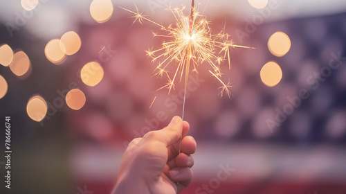 Hand holds sparkler against blurred American flag, celebration and patriotism reflected in bokeh light. Joy of festive spirit glimmers softly photo
