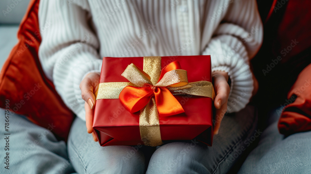 A woman is holding a red box with a gold ribbon on it. The person is sitting on a couch, and the couch is red A woman is holding a red box with a gold ribbon on it. The person is sitting on a couch, and the couch is red