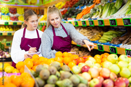 Focused woman grocery store owner giving instructions to young salesgirl, pointing at goods while standing near shelves with fruits and vegetables