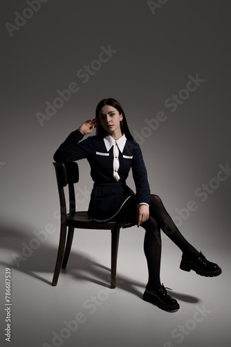Portrait of attractive caucasian young brunette woman in school uniform sitting on chair in studio. Girl posing. Pretty stylish model. Beauty, Style, Fashion, Luxury