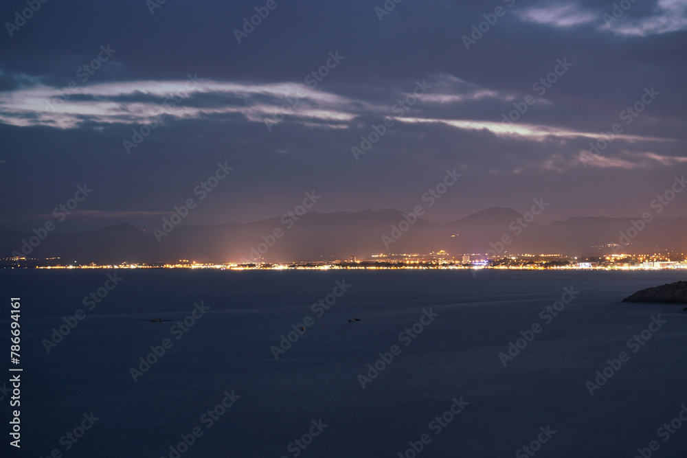  Serene Twilight Over a Coastal City With Glimmering Lights and Distant Mountains. The distant cityscape comes alive with twinkling lights, illuminating the coastline.