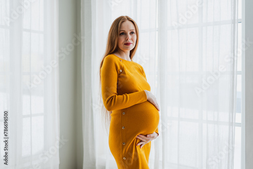 Pregnancy motherhood people expectation future. Pregnant woman with big belly standing near window at home. Girl hugging her tummy enjoying pregnancy. Maternity tenderness parenthood new life concept