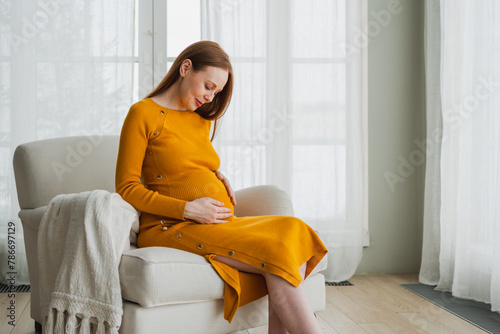 Pregnancy motherhood people expectation future. Pregnant woman with big belly sitting on chair near window at home. Girl hugging her tummy enjoying pregnancy. Maternity tenderness parenthood new life
