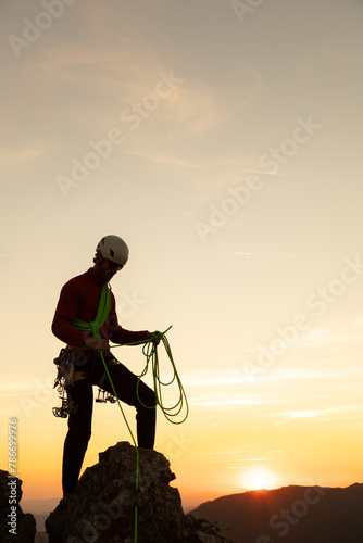A man in a red jacket is standing on a rock with a green rope. The sun is setting in the background, creating a serene and peaceful atmosphere © VICTOR