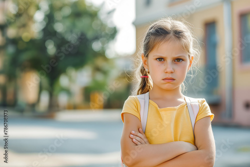 Little grumpy girl standing with her arms crossed in front of school in street. Angry tired child has problem in school. Emotions, stress, learning difficulties