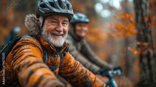 A happy senior couple wearing cycling helmets and jackets, enjoying a bike ride in the autumn forest. The man in focus has a beard and a warm smile. The background features blurred orange foliage.