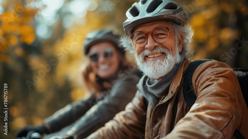 Elderly man and a woman wearing bicycle helmets and jackets, smiling while riding bikes. The man has a beard and glasses, and the background shows autumn foliage. © Vadym Hunko