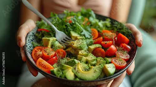 A person is holding a bowl of fresh salad containing avocado slices  cherry tomatoes  lettuce  and herbs. The salad looks healthy and appetizing.