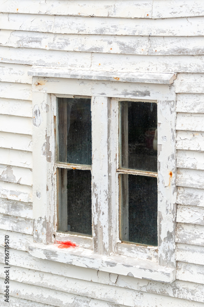 A four pane clear glass window with dirt and grime. The antique window is in a white wooden house with paint peeling. There are rusty nails in the horizontal boards and red paint on the sash. 