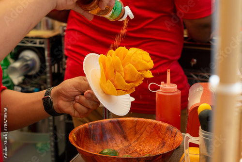 Mexican mango flowers or mango on a stick are made by a street vendor at a market. The fruit is orange color and the vendor is sprinkling lime, chili, salt, and cinnamon over the sticky sweet fruit. 