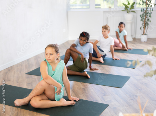 In fitness studio with group of friends, girl children perform Lord of Fishes asana, Ardha Matsyendrasana. Female teen participates in yoga practice. Concept of active lifestyle of youthful generation