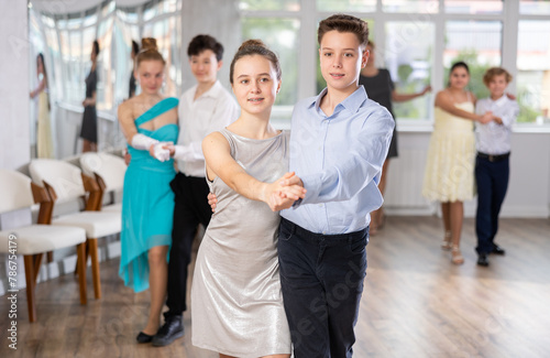 Happy interested teenage students preparing for college festive event, rehearsing ballroom dance steps in spacious classroom while female teacher observing from background..