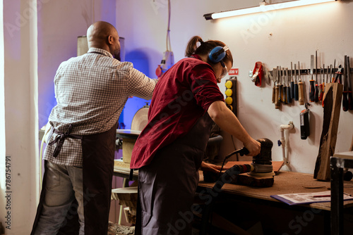 Master carpenter and apprentice using orbital sander on lumber, assembling furniture. Teamworking employees in assembly shop using angle grinder to create wooden decorations, refining wood objects