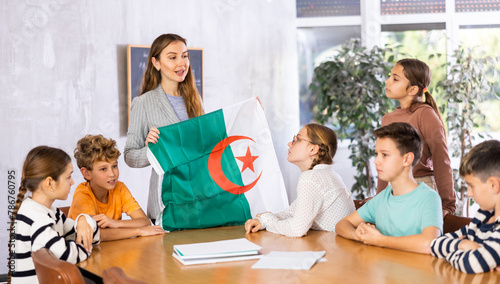 Smiling young woman teacher showing state flag of Algeria and telling preteens schoolchildren history of country during lesson in class